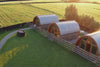 A cinematic aerial view of luxury wooden glamping pods on a farm, each with a private hot tub, spaced across a green field at sunset.